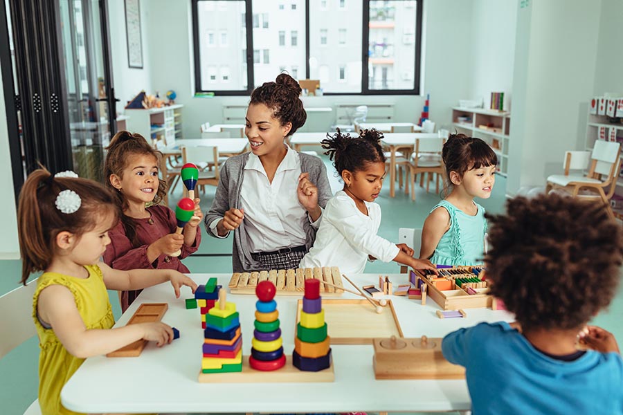 Business Insurance - Preschool Teacher Sings at a White Table as Young Children Play Instruments and Sort Wooden Blocks Around Her