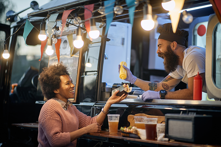 Specialized Business Insurance - Food Truck Owner Leans Out of His Festive Food Truck, Adding Sauce to a Customer’s Sandwich, Both Laughing